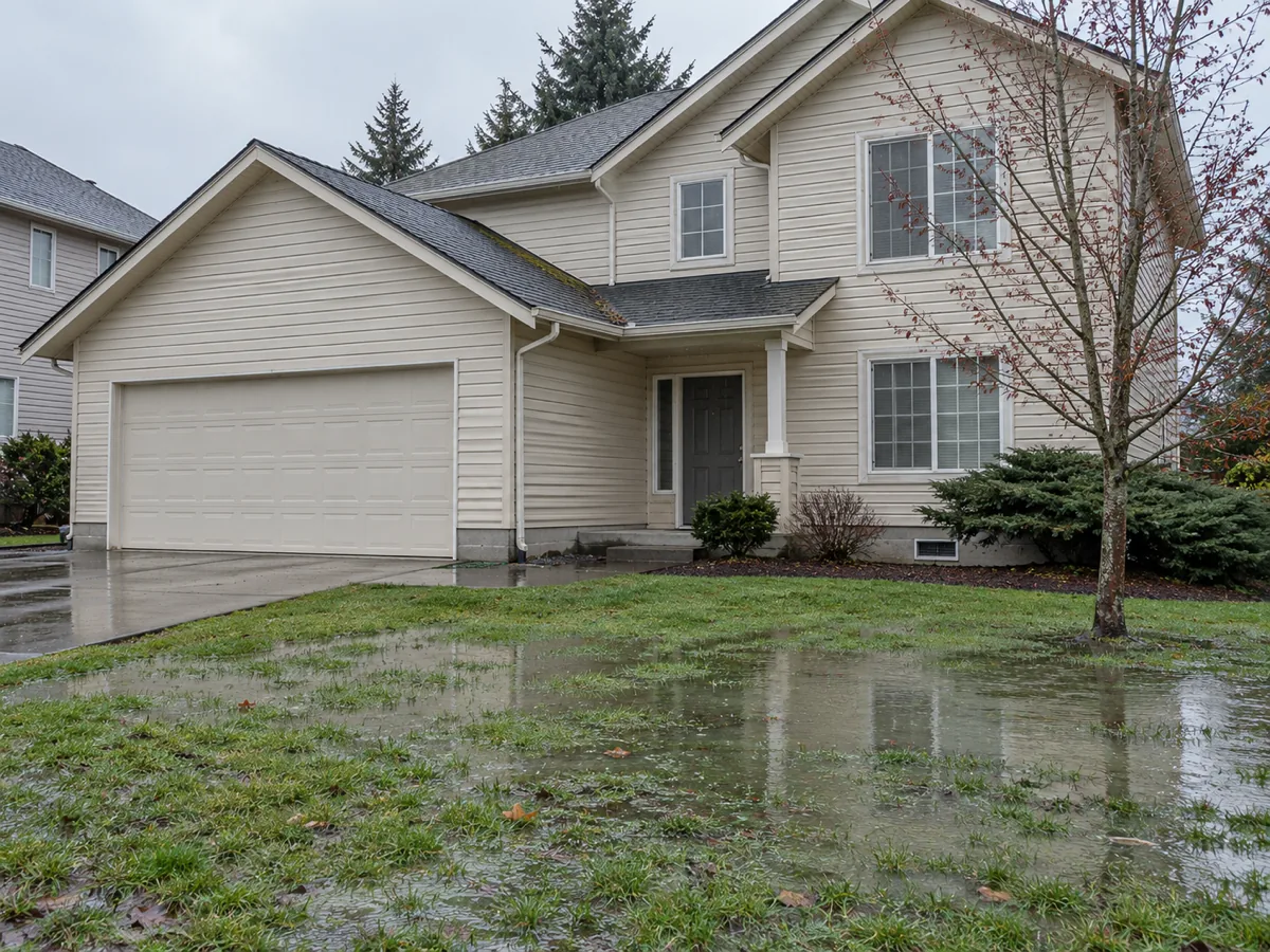 A 1990s two-storey vinyl-sided home with an attached garage and a waterlogged lawn in Auburn, WA