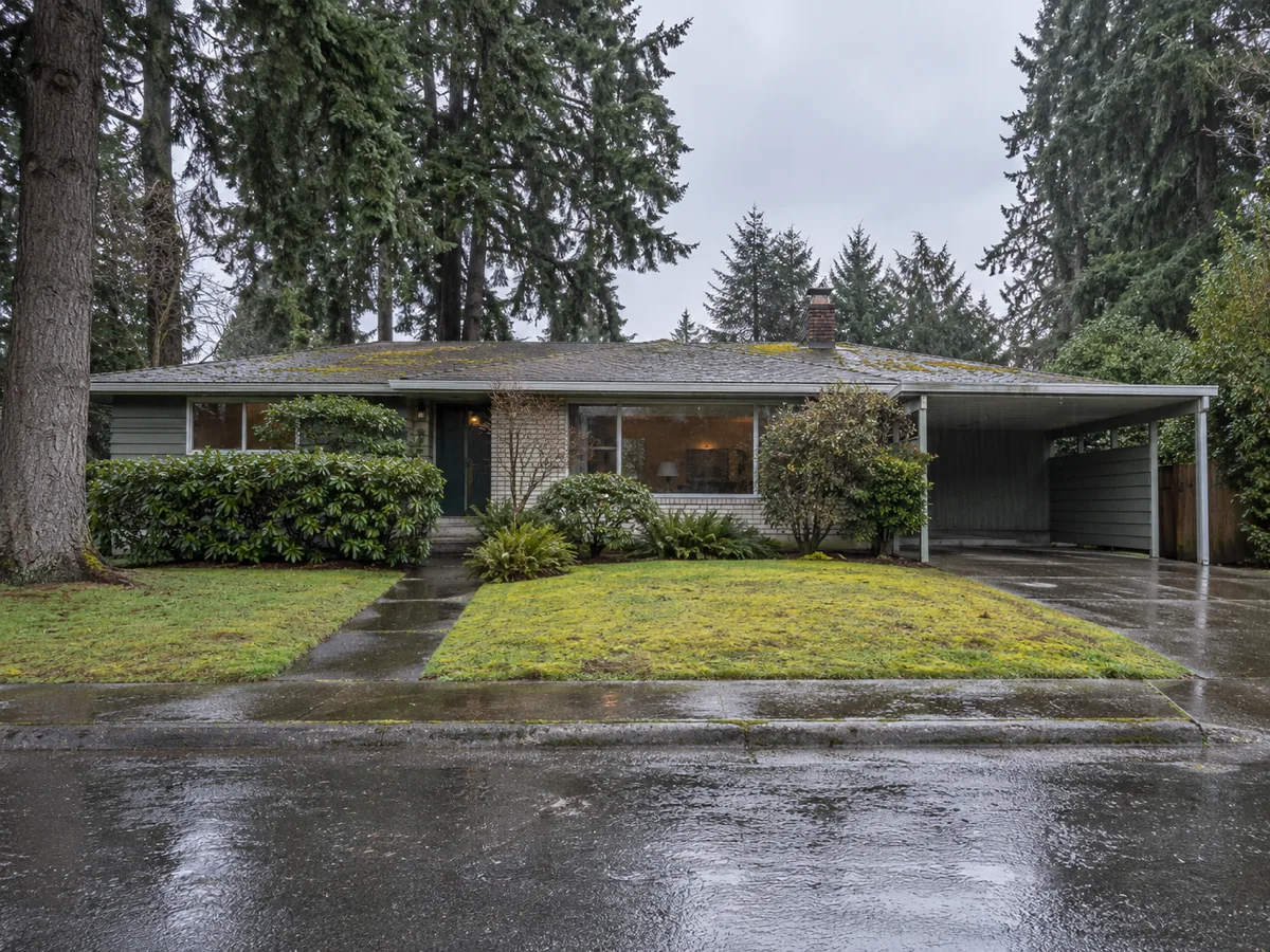 A 1960s single-storey rambler with wide eaves and a carport on a wet street in Burien, WA